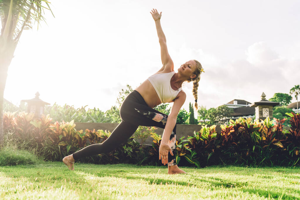 Jovem mulher loira em activewear fazendo exercício de alongamento enquanto pratica ioga no ângulo lateral posar no gramado verde do resort exótico ao nascer do sol - Foto, Imagem