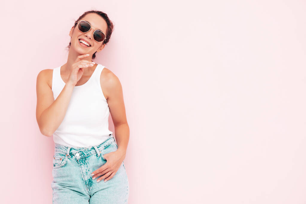 Jonge mooie glimlachende vrouw in witte zomer t-shirt en jeans. Sexy zorgeloze vrouw poseren in de buurt van roze muur in de studio. Positief model dat plezier heeft en gek wordt. Vrolijk en gelukkig. In een zonnebril. - Foto, afbeelding