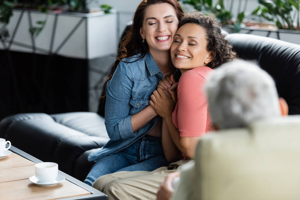 happy multiethnic lesbian women with closed eyes holding hands while sitting on couch near blurred psychologist - Photo, Image