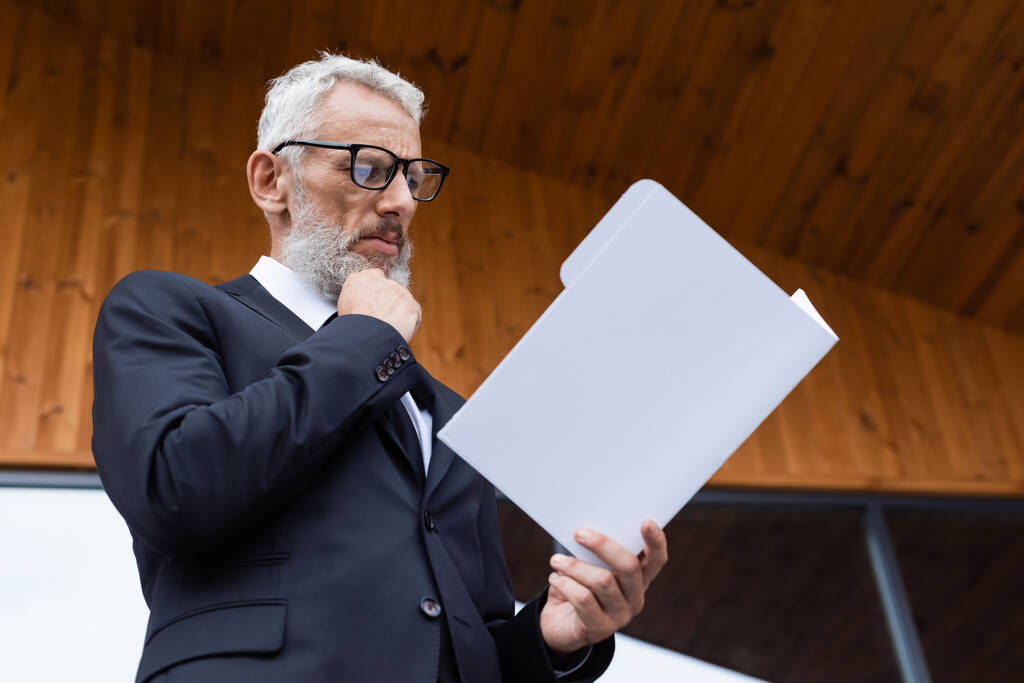 Nachdenklicher Geschäftsmann mittleren Alters mit Brille beim Blick auf Dokumente im Freien - Foto, Bild