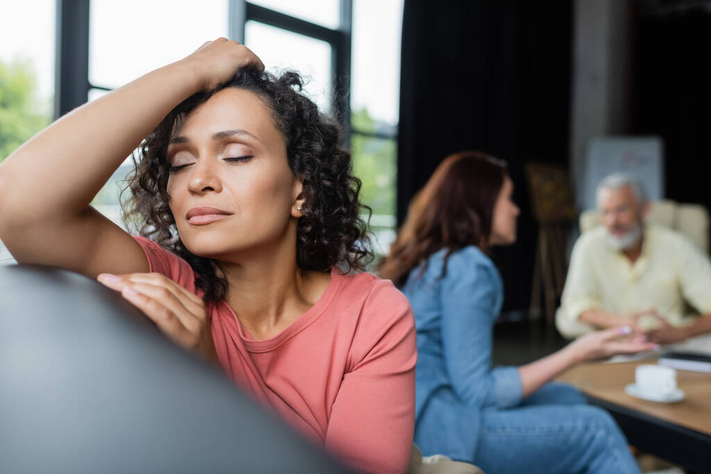 depressieve Afro-Amerikaanse vrouw zitten met gesloten ogen in de buurt van vriendin en psycholoog op wazige achtergrond - Foto, afbeelding