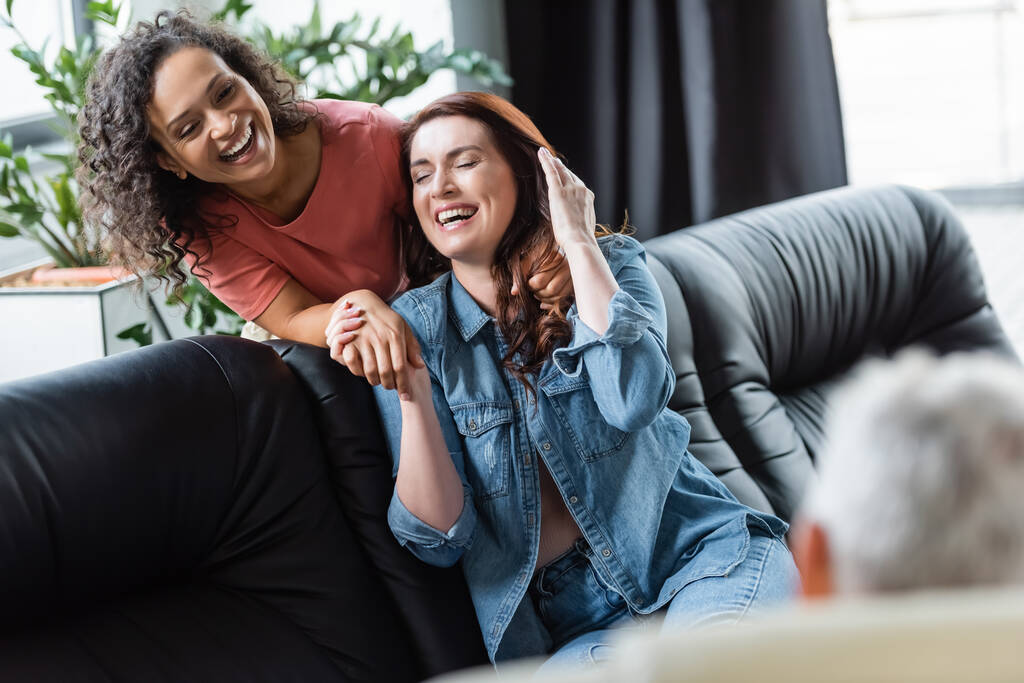 excitada pareja de lesbianas interracial tomados de la mano y riendo cerca de psicólogo borrosa durante la consulta - Foto, Imagen