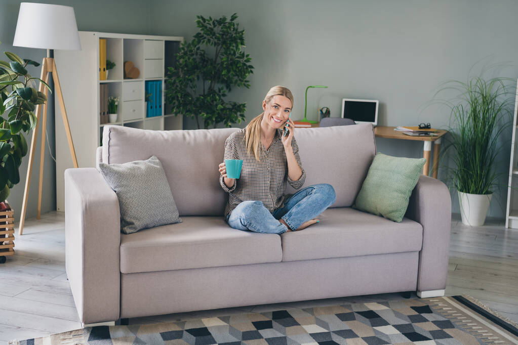 Plná délka fotografie pozitivní dáma sedící divan zkřížené nohy držet cappuccino pohár volání chytrý telefon přítel v obývacím pokoji - Fotografie, Obrázek