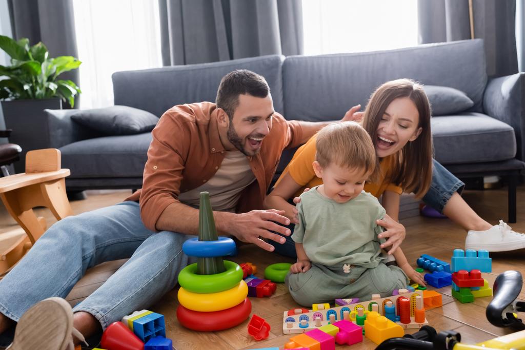 Pais positivos abraçando criança perto de brinquedos e bicicleta na sala de estar  - Foto, Imagem