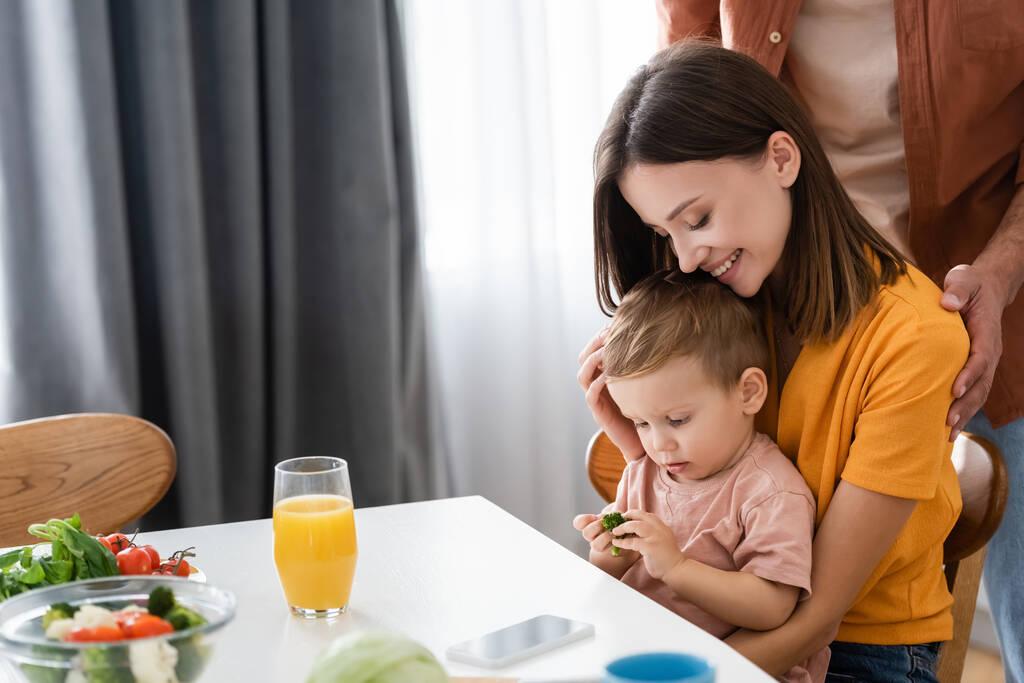Femme gaie étreignant enfant avec brocoli près de mari et salade à la maison  - Photo, image