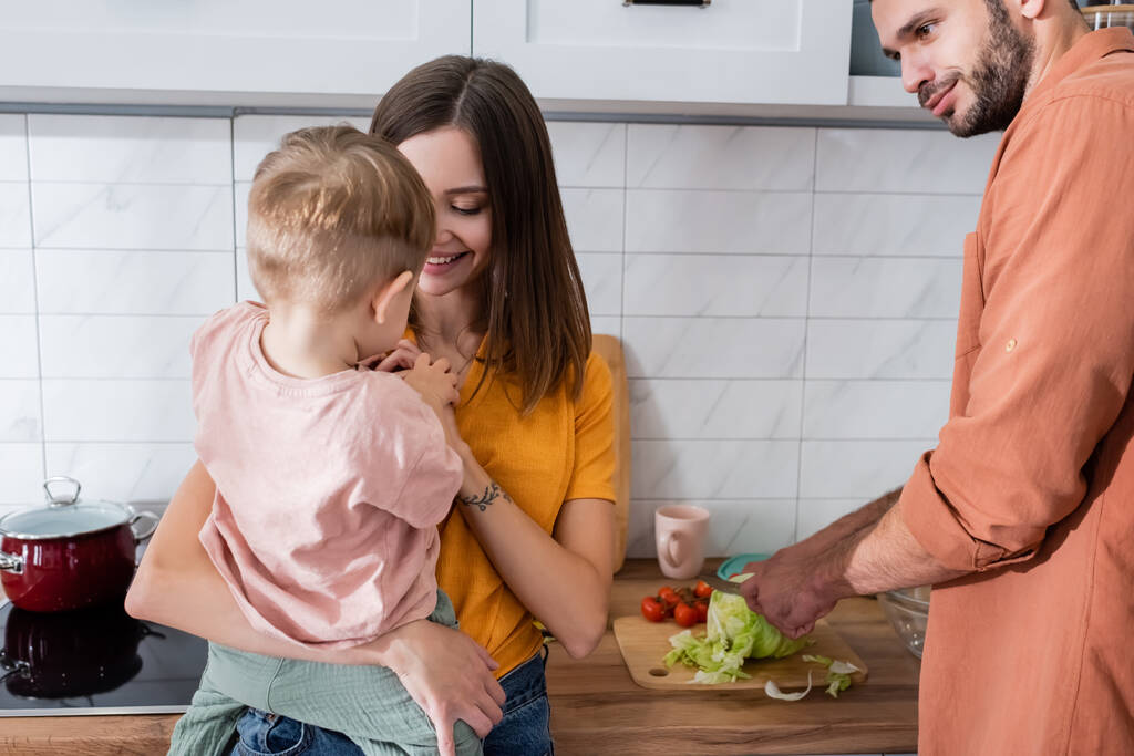 Мужчина готовит салат рядом с улыбающейся женой и ребенком на кухне  - Фото, изображение