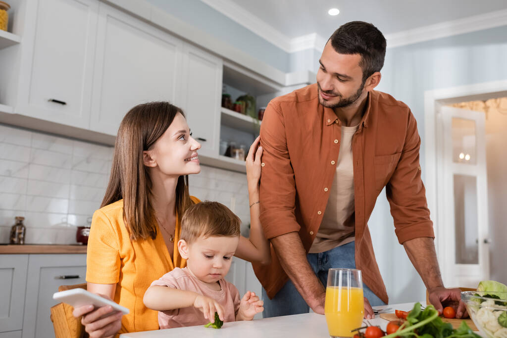 Ευτυχισμένη γυναίκα κρατώντας smartphone κοντά στο γιο και το σύζυγο μαγείρεμα σαλάτα στην κουζίνα  - Φωτογραφία, εικόνα