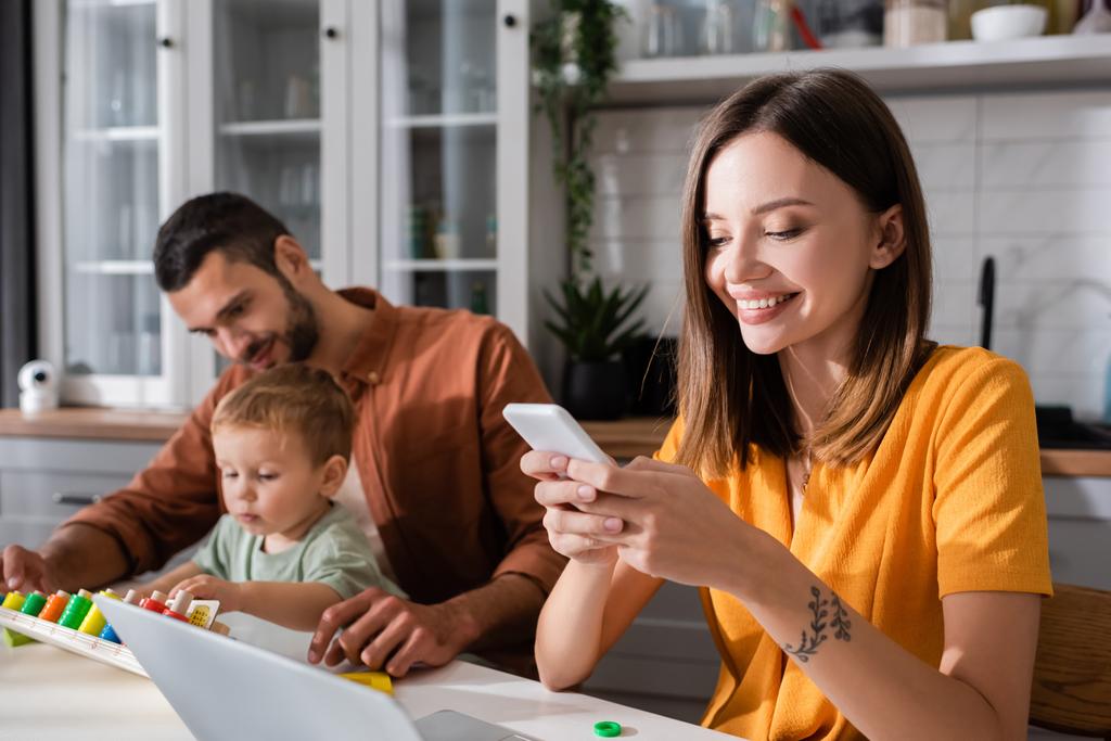 Позитивный фрилансер, использующий смартфон рядом с ноутбуком и семьей, играющий в игры дома  - Фото, изображение