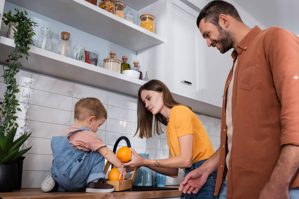 Mujer sosteniendo naranjas cerca de niño y marido en la cocina  - Foto, Imagen