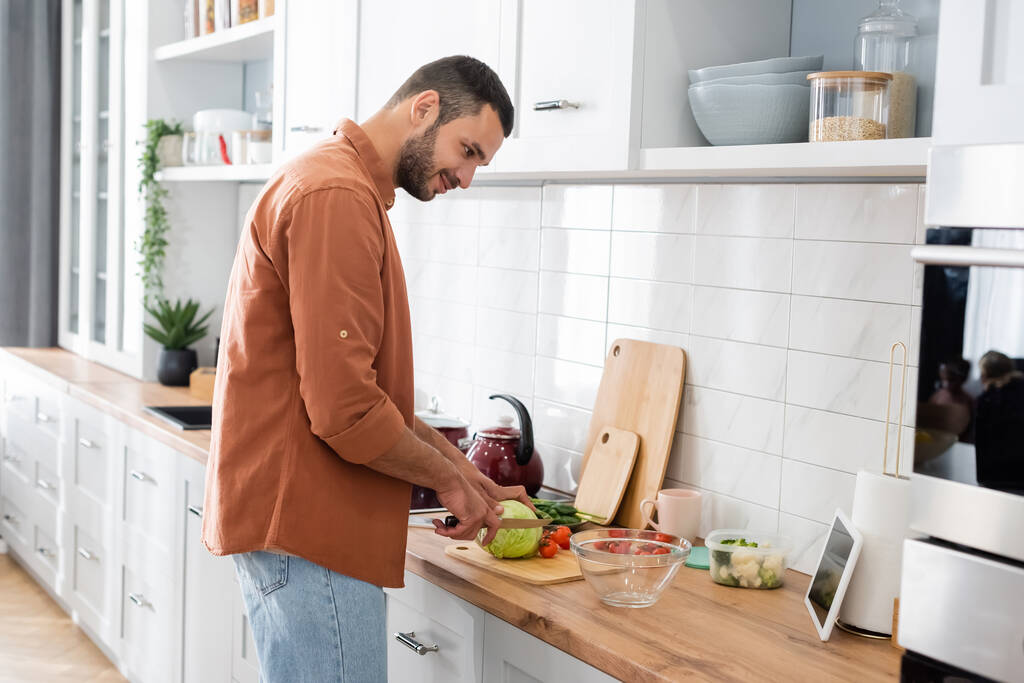 Vista lateral del hombre sonriente mirando tableta digital mientras corta verduras en la cocina  - Foto, imagen