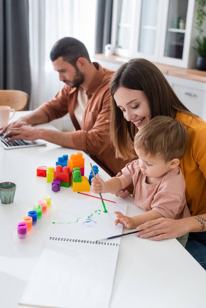 Mosolygós nő festés fiával közel építőkockák és férj segítségével laptop otthon  - Fotó, kép