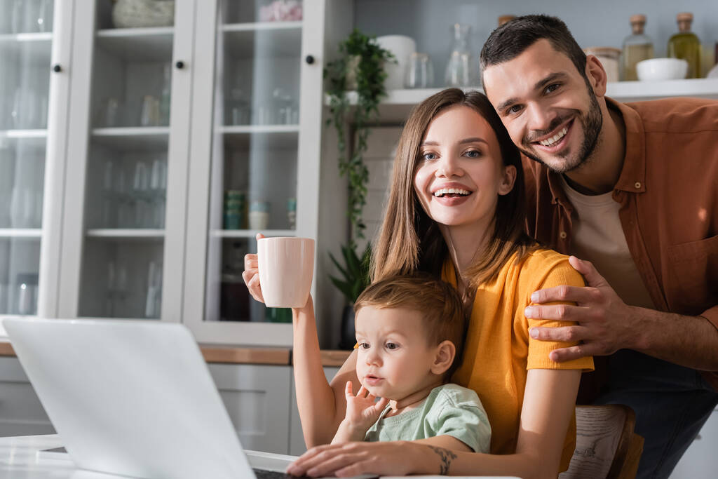 Uśmiechnięta rodzina patrząca na kamerę w pobliżu syna i laptopa w domu  - Zdjęcie, obraz
