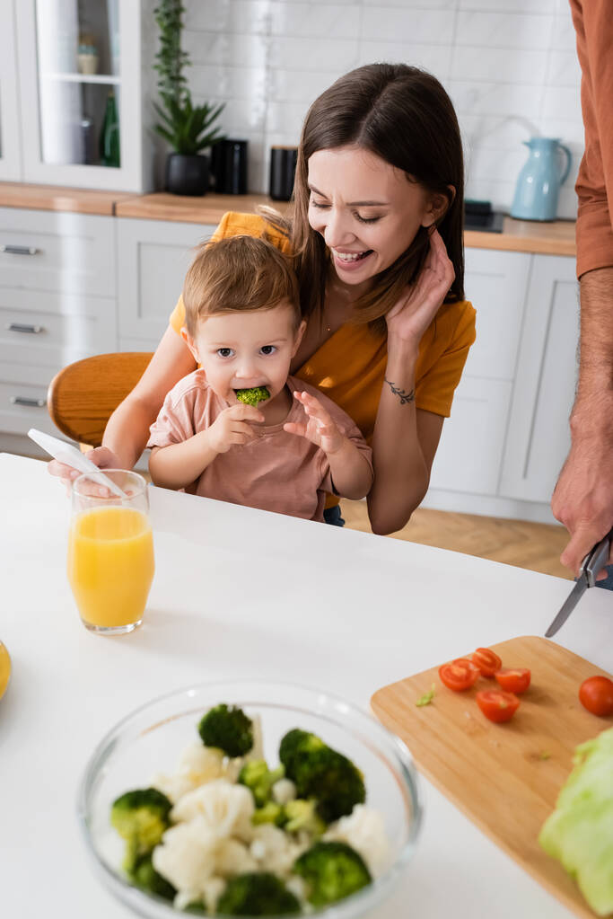 Happy mother holding smartphone and looking at child eating broccoli at home  - Photo, Image