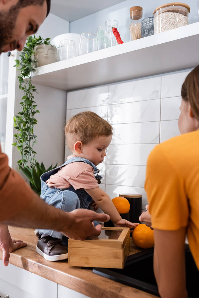 Niño sosteniendo naranjas cerca de los padres en la cocina  - Foto, Imagen