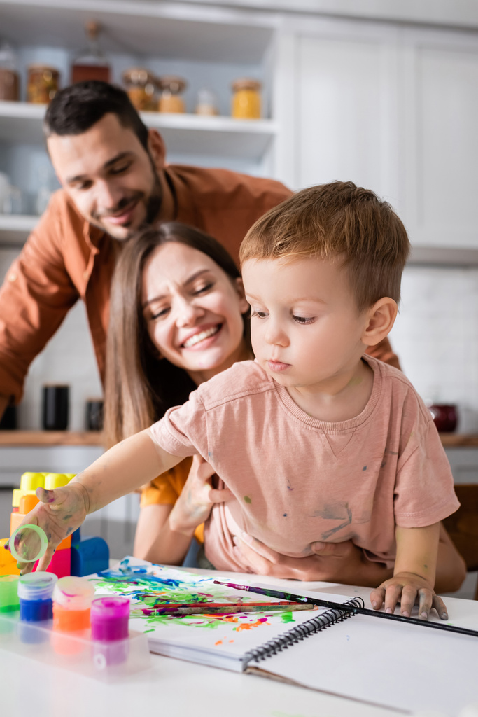 Toddler gyerek játszik festék közelében vázlatfüzet és elmosódott szülők otthon  - Fotó, kép