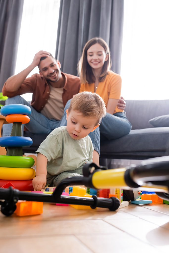 Bambino bambino seduto vicino a giocattoli e bici vicino ai genitori in soggiorno  - Foto, immagini