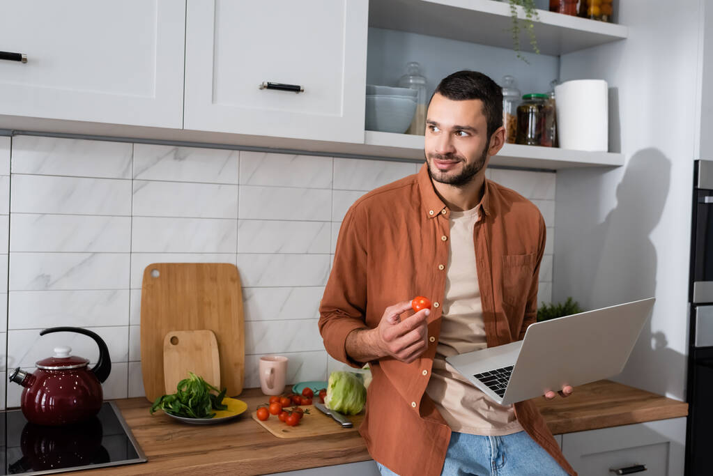 Homme souriant tenant ordinateur portable et tomate cerise dans la cuisine  - Photo, image