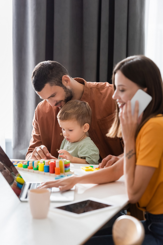 Sonriente hombre sentado cerca de hijo jugando juego educativo y esposa trabajando en el ordenador portátil y hablando en el teléfono celular en casa  - Foto, imagen