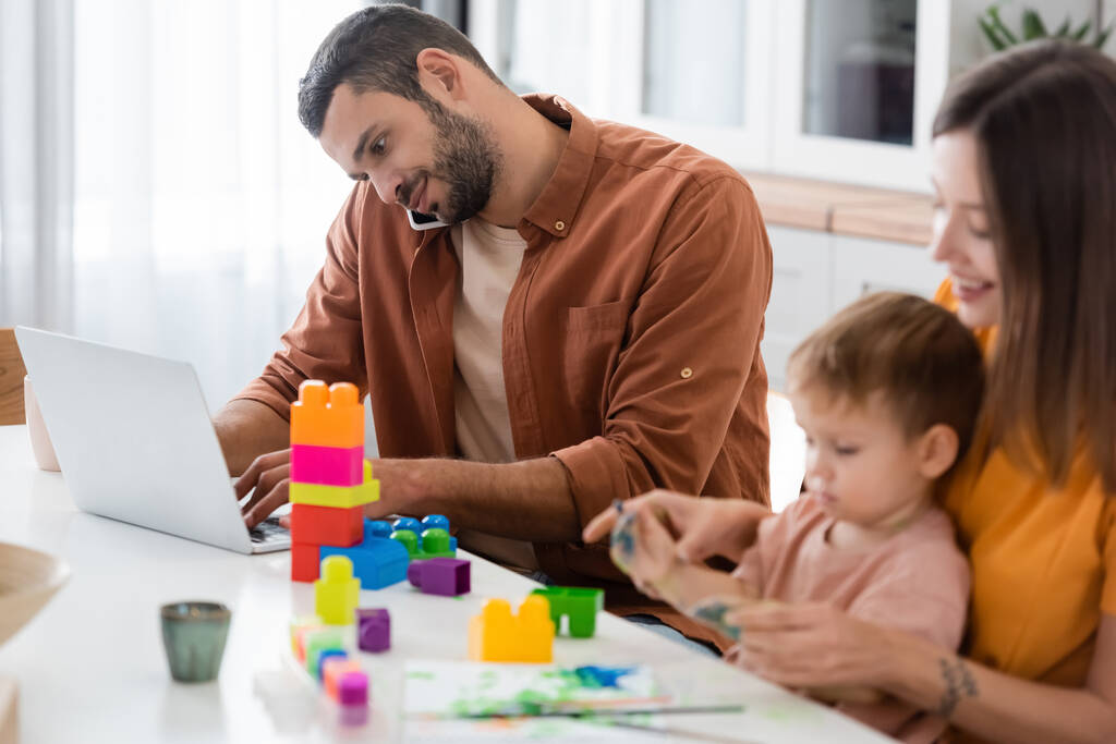 Man using laptop and talking on smartphone near family with building blocks at home  - Photo, Image