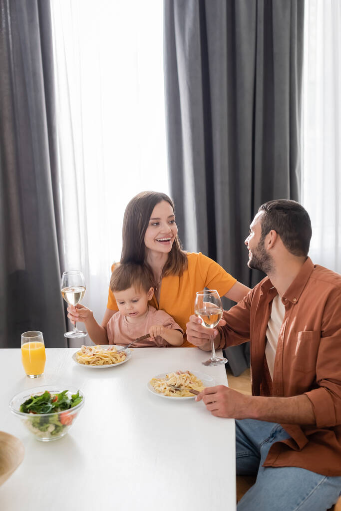 Χαρούμενη γυναίκα που κρατά το κρασί κοντά στο σύζυγο και το γιο κατά τη διάρκεια του δείπνου στο σπίτι  - Φωτογραφία, εικόνα