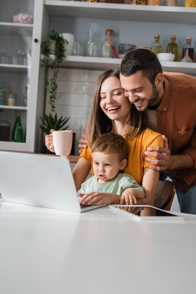 Cheerful husband hugging wife with cup near gadgets and son at home  - Photo, Image