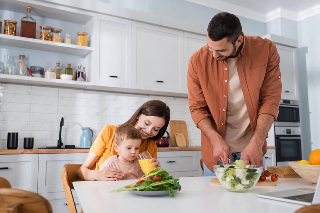 Mujer feliz sosteniendo jugo de naranja cerca de hijo y marido cortando verduras en la cocina  - Foto, imagen