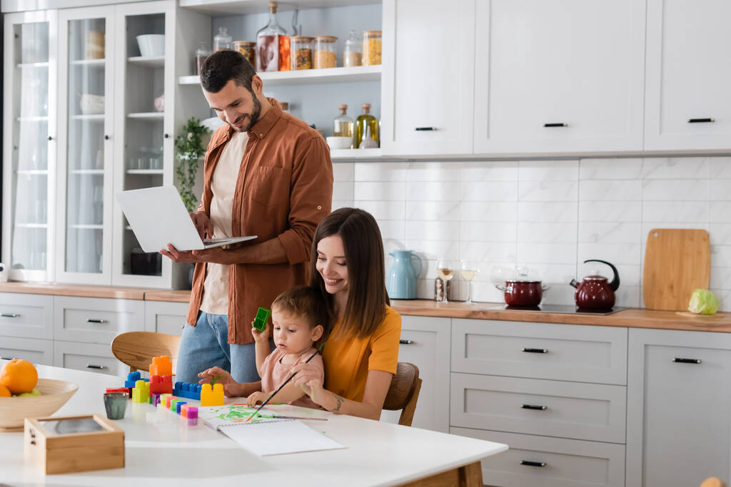 Homme utilisant un ordinateur portable et regardant le dessin de la famille dans la cuisine  - Photo, image
