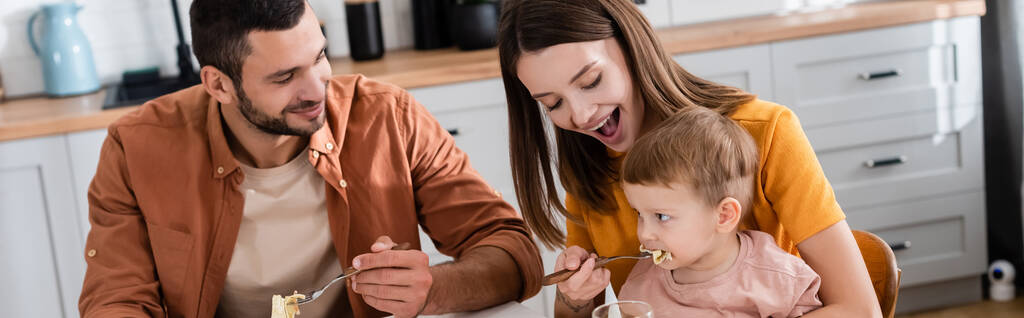 Jovem mulher alimentando filho com macarrão perto do marido em casa, banner  - Foto, Imagem