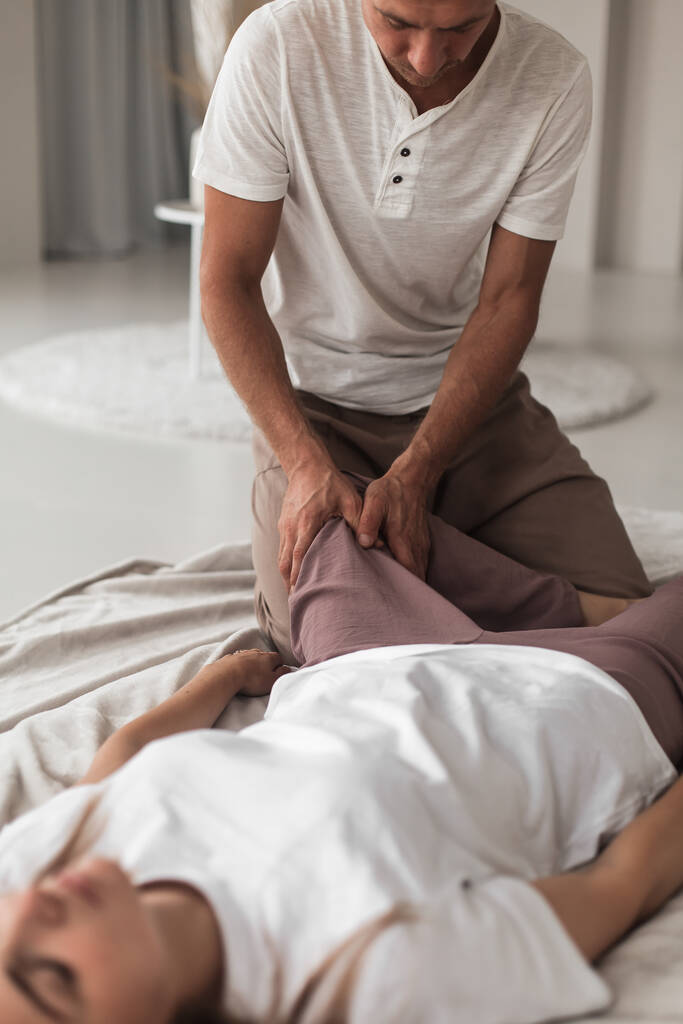 Egy profi fiatalember masszírozza egy nő lábát egy fehér, hangulatos szobában egy manuális terápia alatt. - Fotó, kép