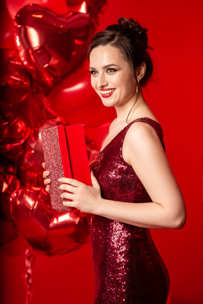 Krásná mladá žena v červených večerních šatech pózuje nad červeným pozadím s velkými balónky ve tvaru srdce. - Fotografie, Obrázek