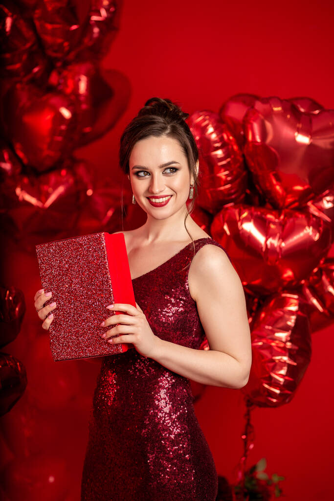 Schöne junge Frau in rotem Abendkleid posiert vor rotem Hintergrund mit großen herzförmigen Luftballons. - Foto, Bild