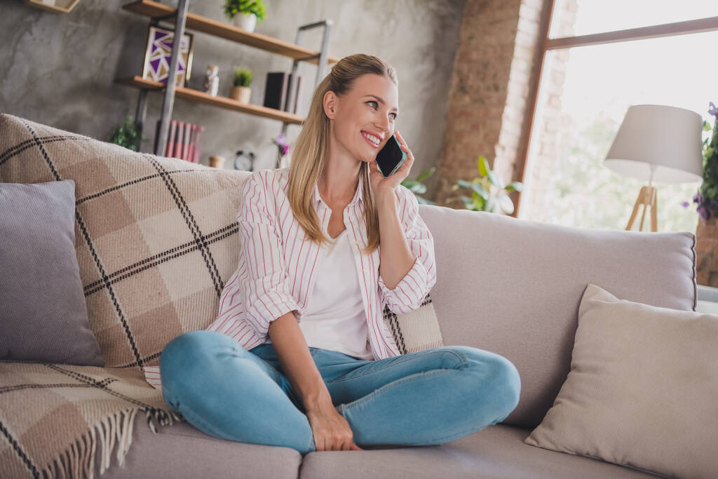 Полноразмерное фото очаровательной зрелой блондинки, сидящей на диване, разговаривающей по телефону, одетой в джинсы в белой рубашке дома в карантине - Фото, изображение