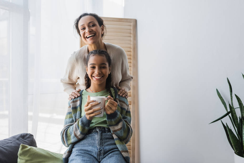 vreugdevolle Afrikaan amerikaanse vrouw en tiener meisje met kopje thee glimlachen op camera thuis - Foto, afbeelding