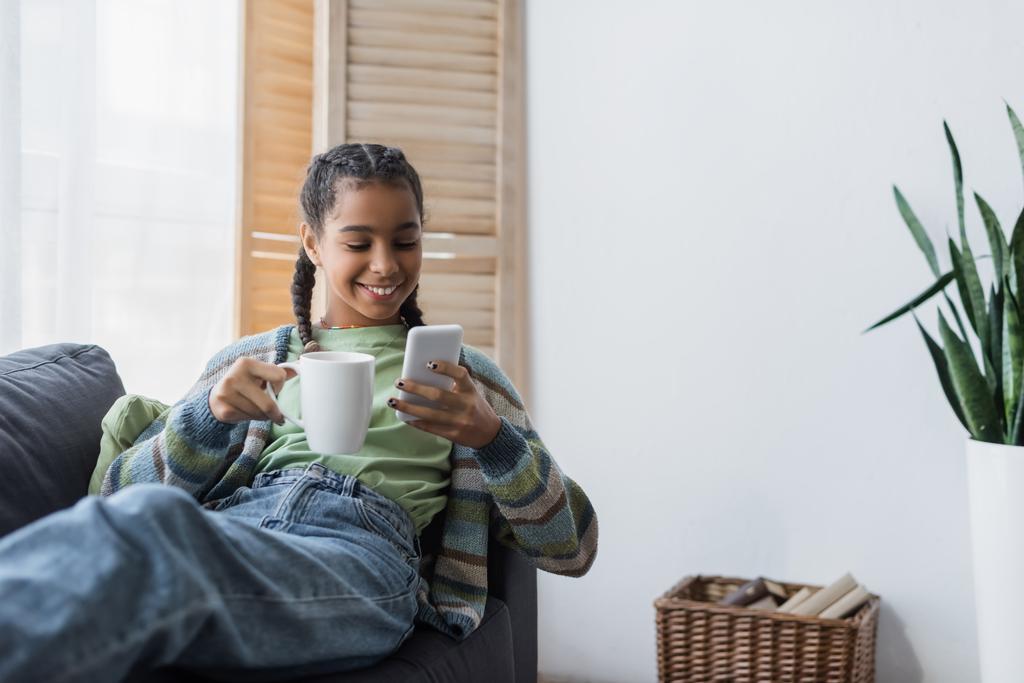 mosolygós afro-amerikai tinédzser lány csevegés mobiltelefon, miközben kezében csésze teát - Fotó, kép