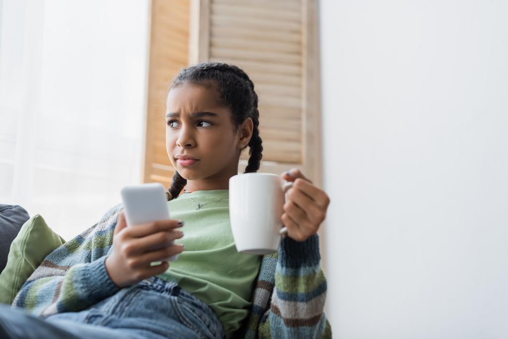 malestar adolescente afroamericano sosteniendo taza de té y teléfono móvil borroso en casa - Foto, Imagen