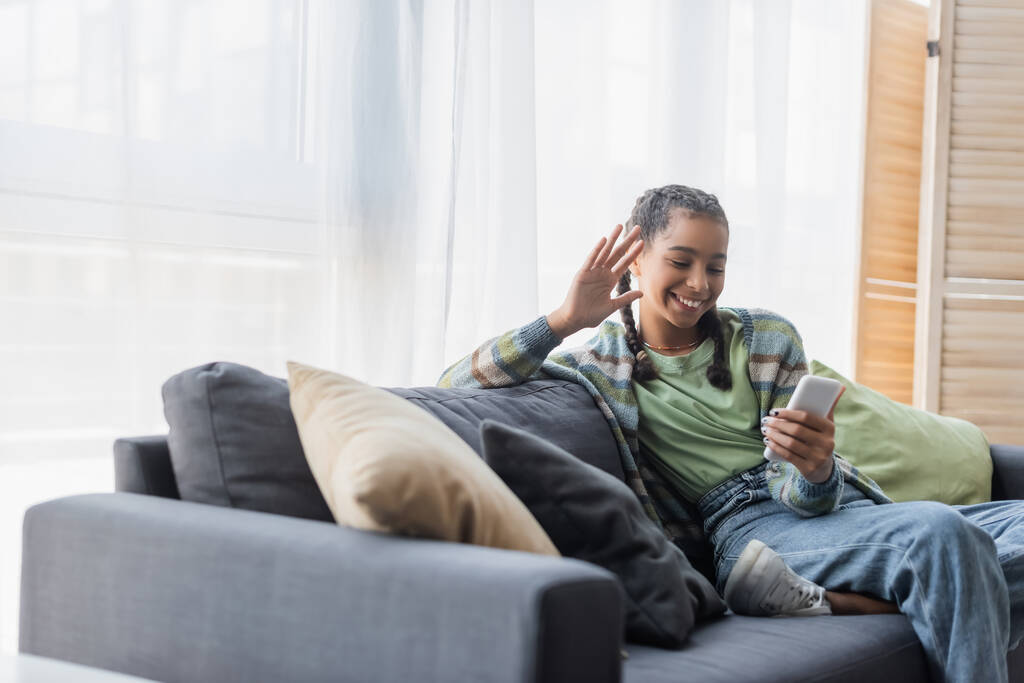 vrolijk Afrikaans amerikaans tiener meisje zitten op de bank met kussens en zwaaien hand tijdens video call op smartphone - Foto, afbeelding