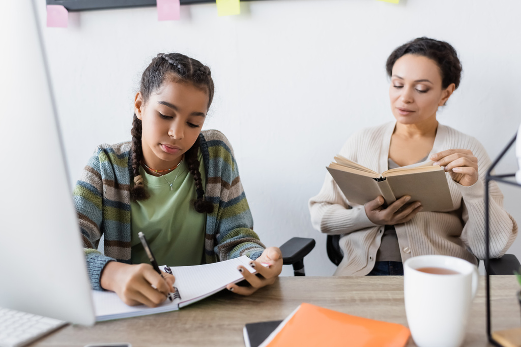 afro-américaine adolescent fille écriture dans notebook près flou ordinateur et maman livre de lecture - Photo, image