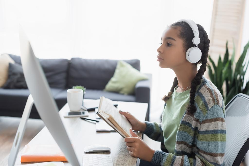 афроамериканська дівчина-підліток в навушниках, дивлячись на монітор комп'ютера, тримаючи книгу
 - Фото, зображення