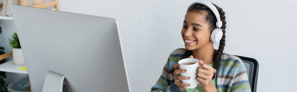 joyful african american schoolgirl in headphones holding cup of tea near computer monitor, banner - Photo, Image