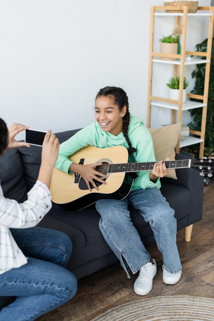woman taking photo of teenage african american daughter playing guitar on couch - Photo, Image