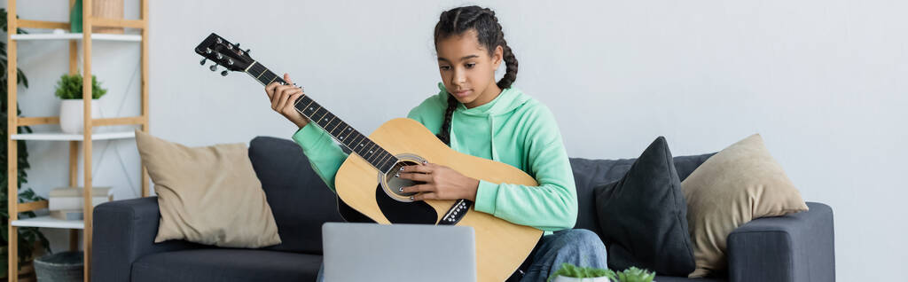 africana americana adolescente menina sentado no sofá com travesseiros e aprender a tocar guitarra perto de laptop, banner - Foto, Imagem