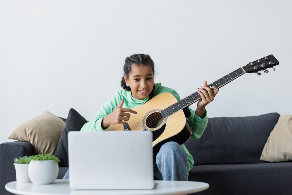 uśmiechnięta Afroamerykanka patrząca na laptopa i wskazująca gitarę ucząc się grać w domu - Zdjęcie, obraz