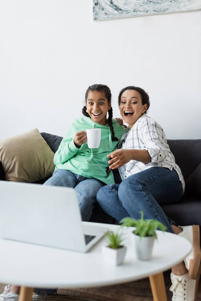 eccitata donna africana americana ridendo e indicando il computer portatile mentre guardava film vicino a figlia adolescente - Foto, immagini
