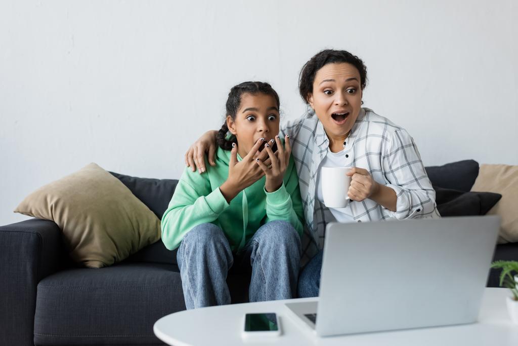 shocked african american woman with teenage daughter watching movie on blurred laptop - Photo, Image