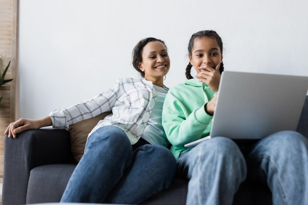 asombrado afroamericano adolescente cubriendo la boca con la mano mientras ve la película en el ordenador portátil cerca de mamá sonriente - Foto, Imagen