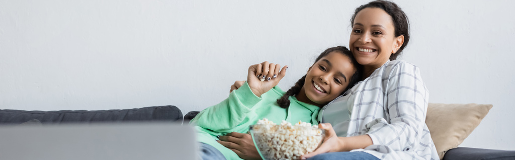 alegre afroamericana mujer y adolescente chica viendo película en portátil cerca de tazón de palomitas de maíz, bandera - Foto, Imagen