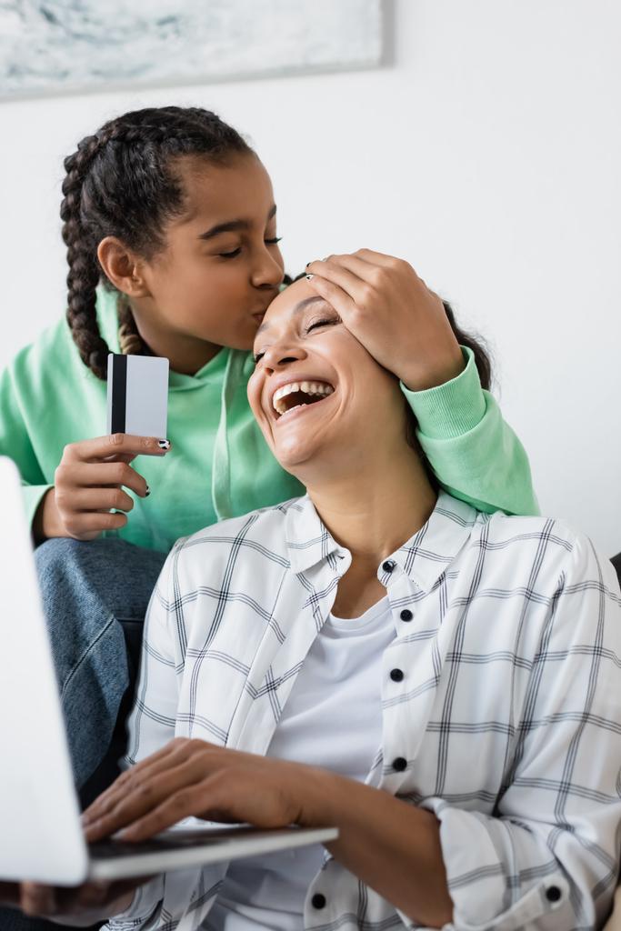 Afrikaans amerikaans meisje zoenen voorhoofd van vrolijke moeder terwijl het houden van credit card in de buurt wazig laptop - Foto, afbeelding