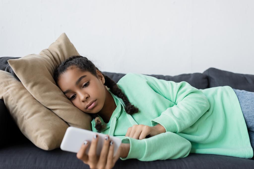 засмучена афроамериканська дівчина-підліток, що використовує мобільний телефон, лежачи на дивані
 - Фото, зображення
