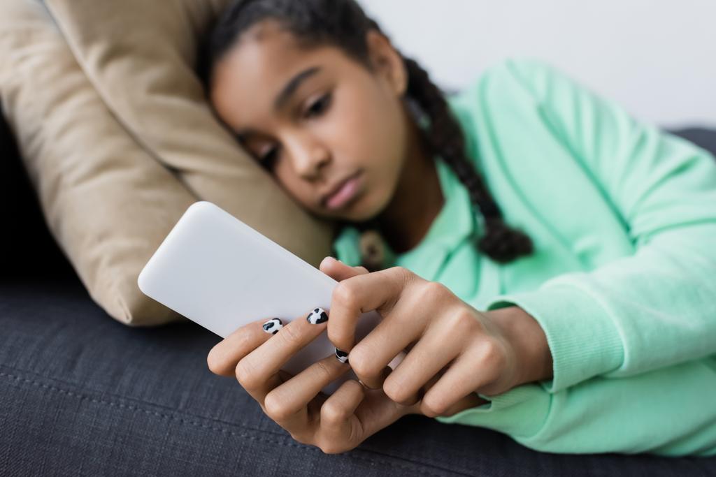 flou afro-américain adolescent fille de mauvaise humeur messagerie sur téléphone mobile tout en étant couché sur le canapé - Photo, image