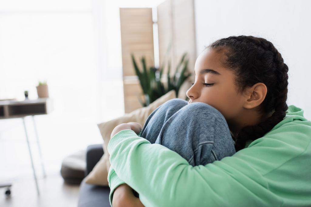 rozrušený africký americký dospívající dívka objímání kolena zatímco sedí se zavřenýma očima doma - Fotografie, Obrázek